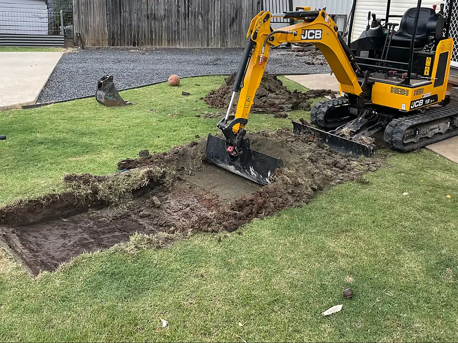 Yellow mini excavator digging a trench through a backyard lawn beside a Queenslander-style house